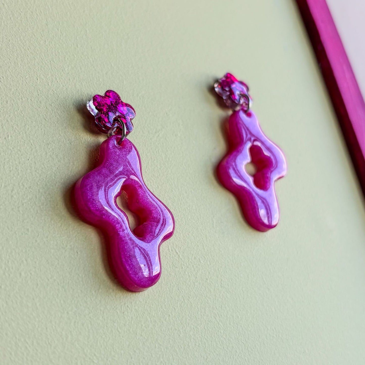 Pendientes de dos piezas, una inferior con forma asimétrica ondulada y una pieza superior pequeña en forma de flor que va pegada al lóbulo de la oreja. Color rosa brillante