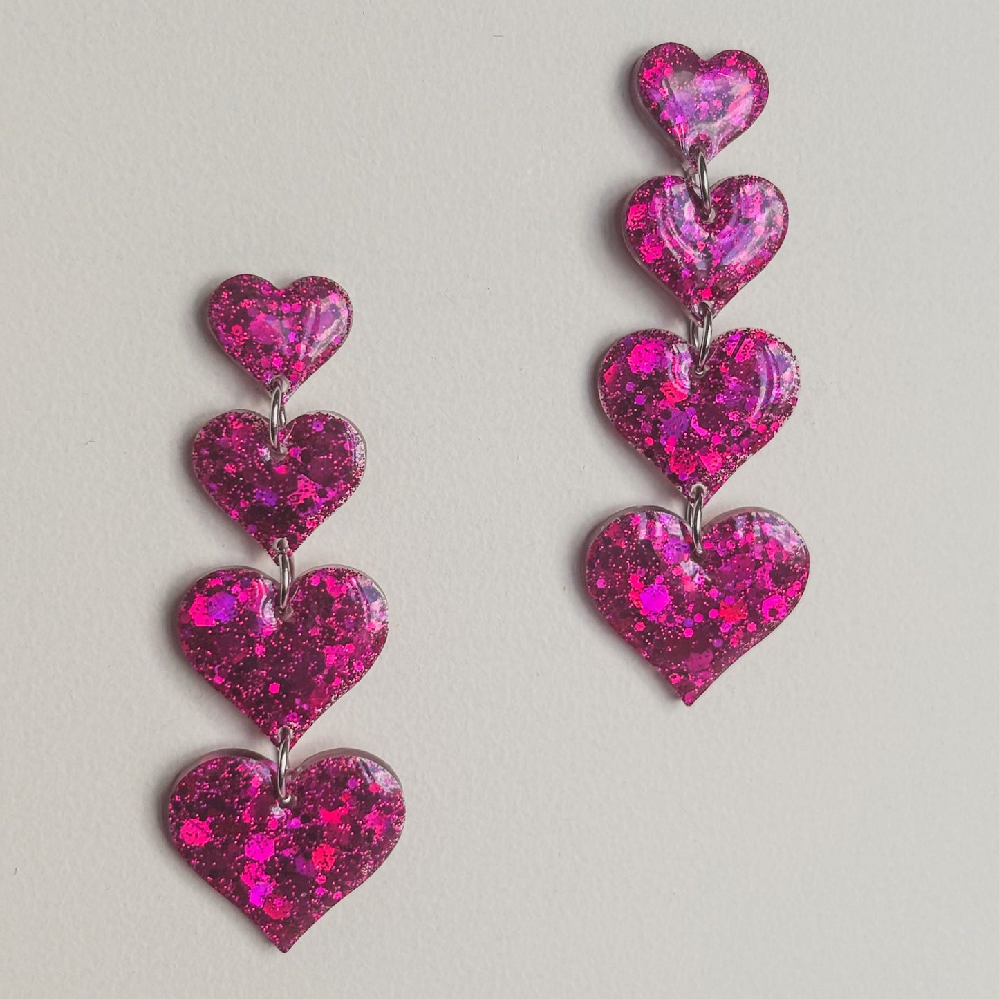 Pendientes artesanales de resina epoxi y acero quirúrgico hipoalergénico. Forma de corazones en cascada color fucsia con mucho brillo