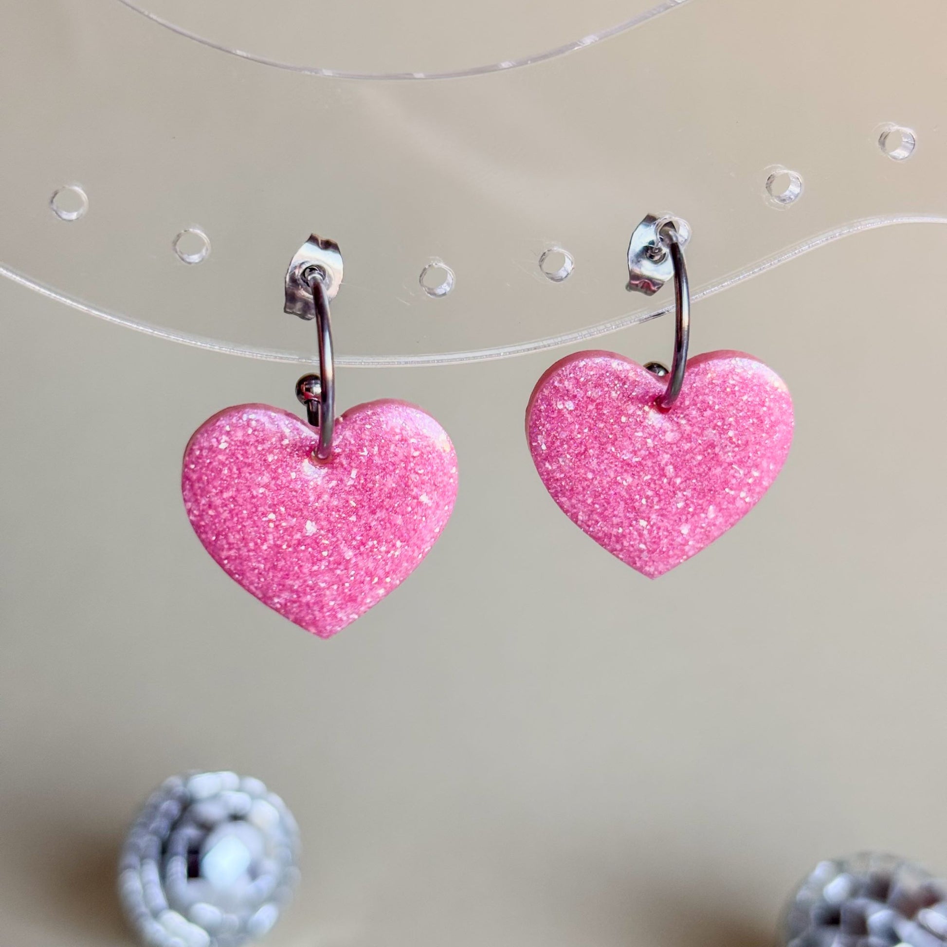 Expositor transparente con pendientes de corazones rosa con brillo metalizado y aros plateados hipoalergénicos.